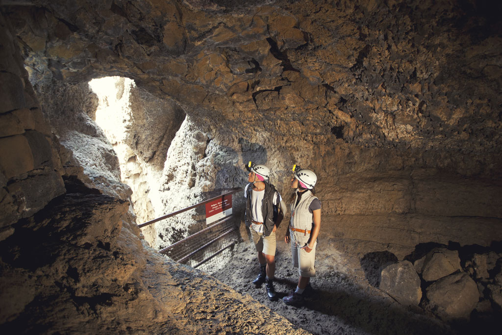 Two people wearing helmets explore a cave in Tenerife, with natural light streaming in.