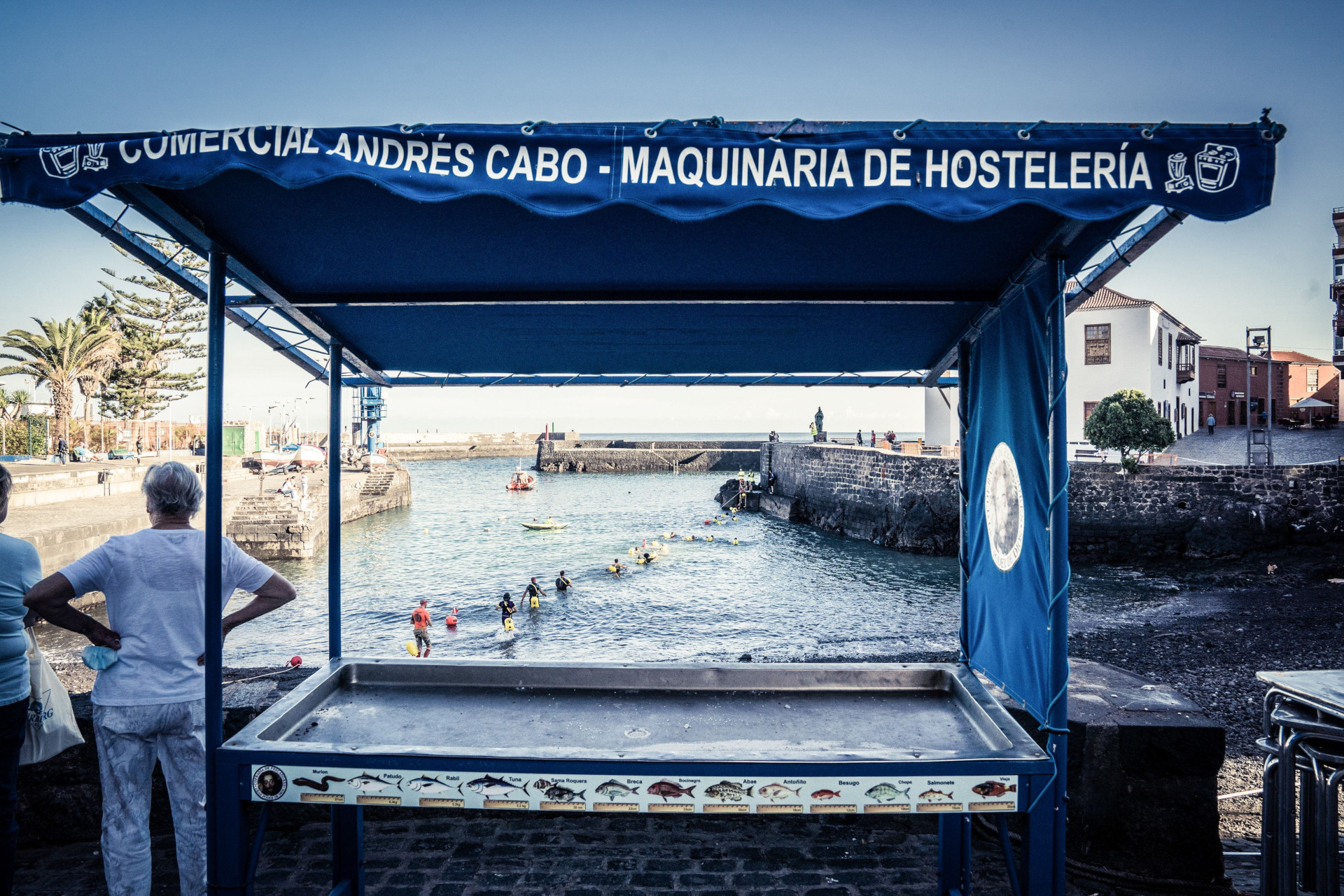 Empty stall by the sea in Tenerife, with people swimming in the background.