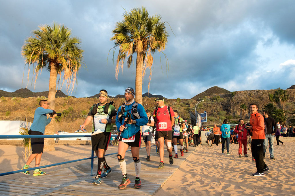 Runners walk along the beach in San Andrés, Tenerife, with palm trees in the background.