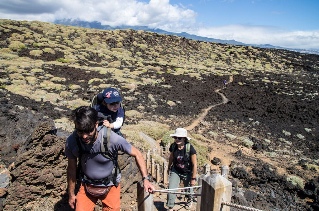 People walking along the volcanic trail in Tenerife under partly cloudy skies.