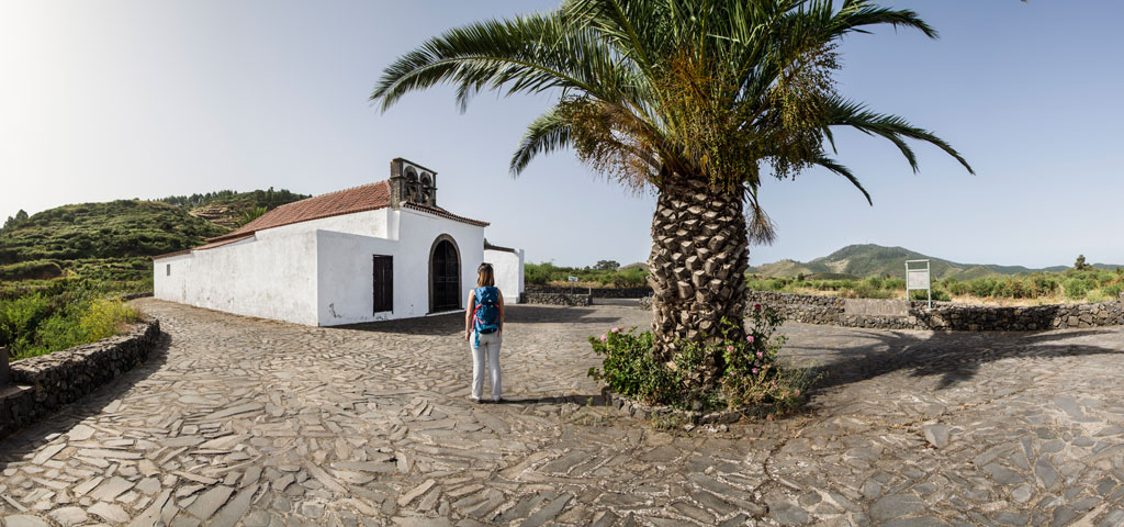 A person with a backpack observes the hermitage surrounded by vegetation in Tenerife.