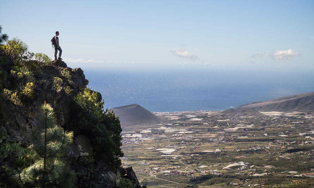 Person contemplating the landscape from a peak in Tenerife, overlooking the ocean and fields.