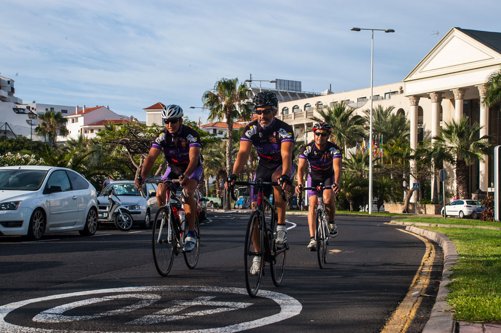 Cyclists walk along a street in Costa Adeje, in the background palm trees and a building.