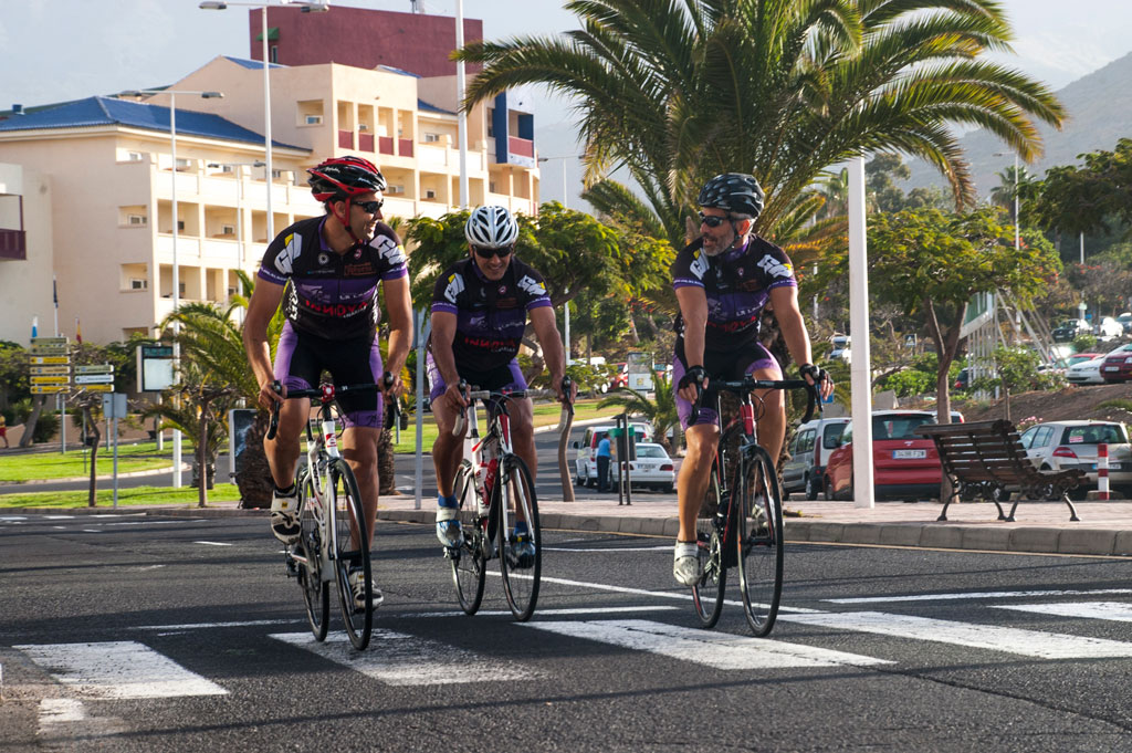 Cyclists cross a road in Tenerife with a building in the background and palm trees around it.