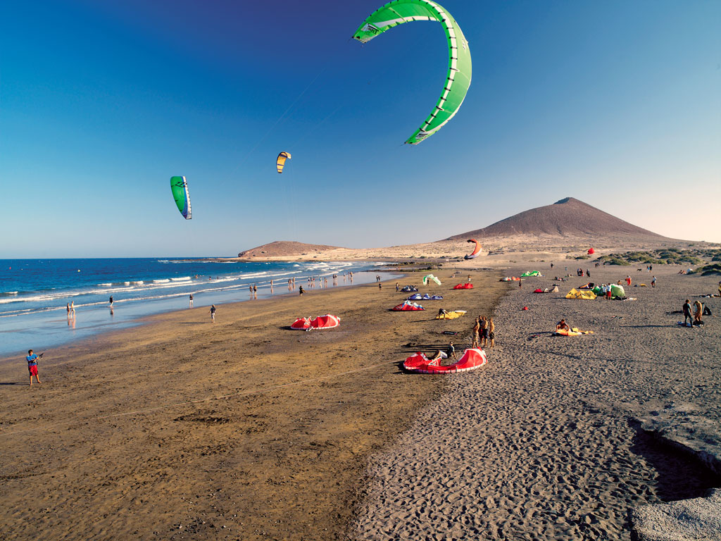 People kitesurfing on the beach of El Médano, Tenerife, with mountains in the background.