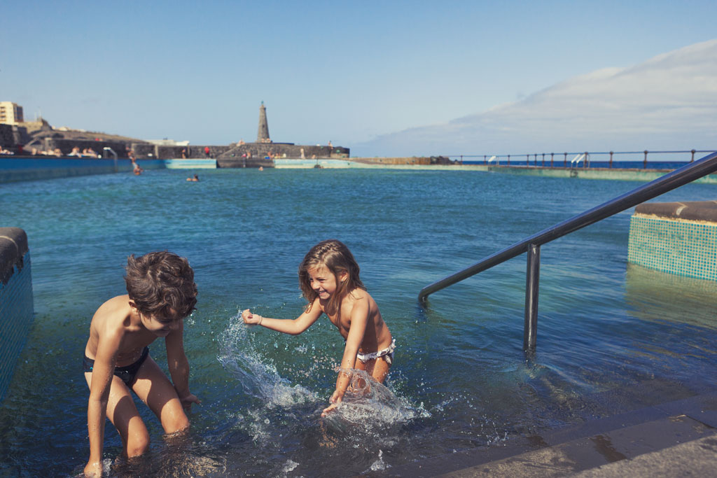 Children playing in a natural pool in Bajamar, Tenerife, with a lighthouse in the background.