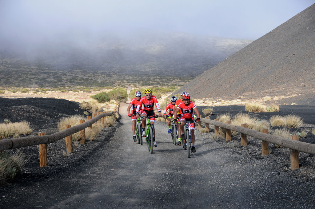 Cyclists ride a volcanic trail in Tenerife under cloudy skies.