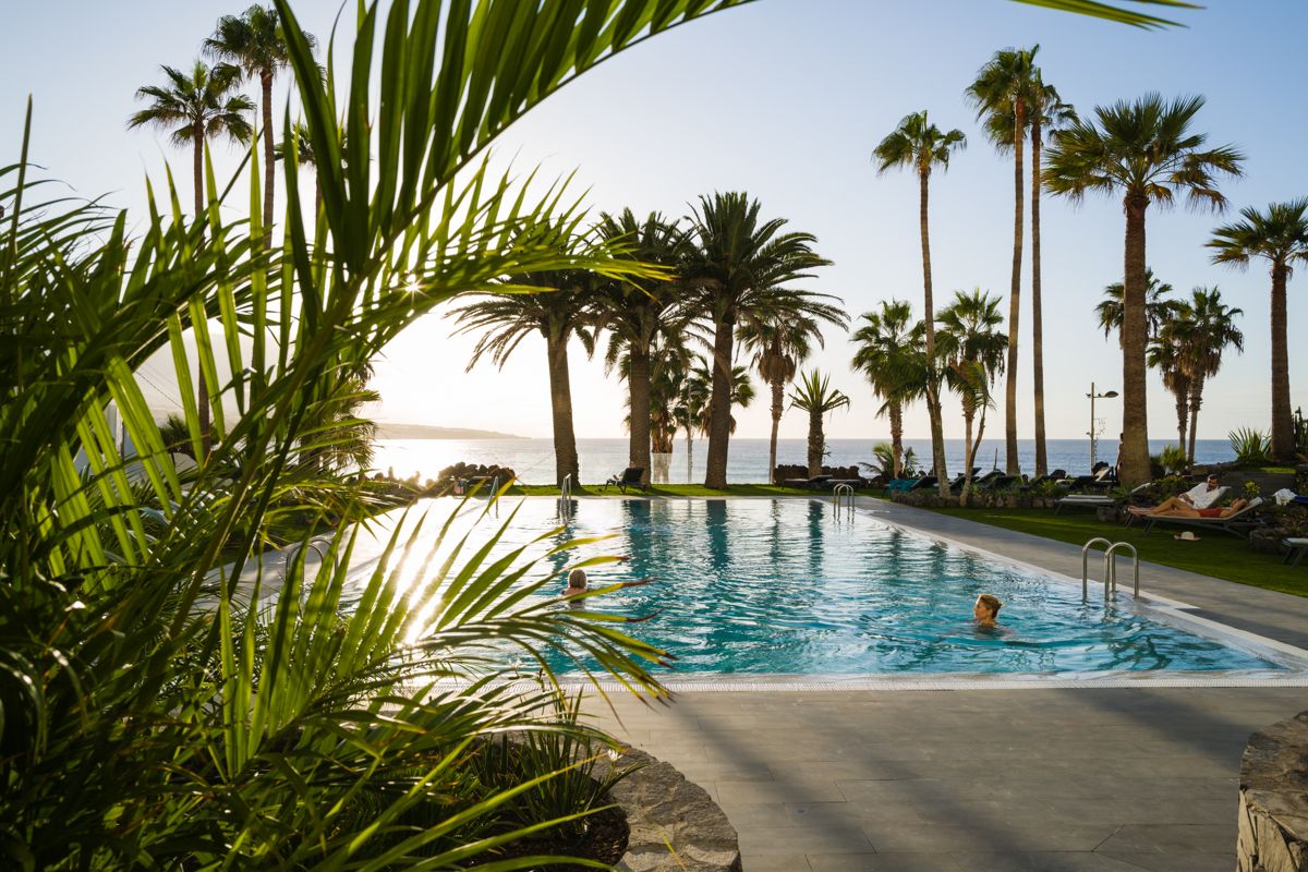 Swimming pool surrounded by palm trees at sunset in Tenerife, overlooking the ocean.